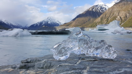 This photo is taken in Mount Cook Glacier in Zealand. A hand stretches out to hold a piece of ice. The ice is crystal clear. It looks very beautiful. In the background is stunning snow mountain.