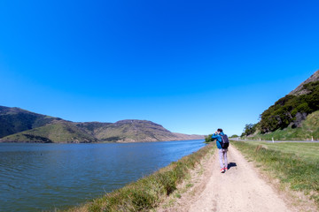 A traveler admiring the beautiful landscape of New Zealand. It has clear blue sky, blue lake, and beautiful mountains and a straight gravel path. This place is peaceful, calm and breathtaking.