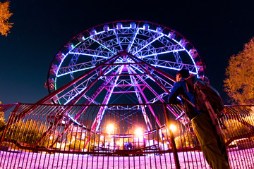 man stands in front of a glowing illumination by a ferris wheel, reflected in a puddle