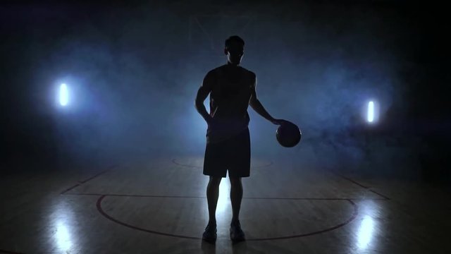 The basketball player goes to the camera and knocks the ball on the ground then stops and holds the ball looking at the camera