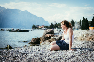 Naklejka na ściany i meble Beautiful young woman is sitting on the pebble beach and enjoying the view, vacation