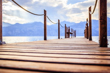 Obraz premium Wooden dock pier extending over blue lake water, mountains at lago di garda