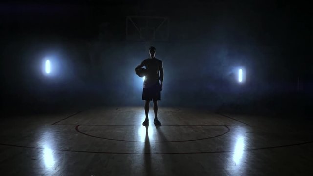 The basketball player stands on a dark Playground and holds the ball in his hands and looks into the camera in the dark with a backlit in slow motion and around smoke