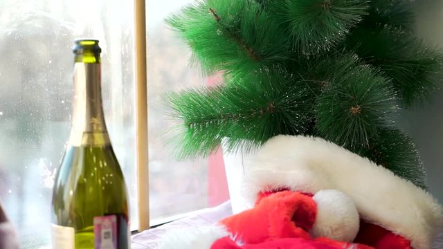 Santa Claus hat on white sill against the window and artificial spruce branches. Red hat, opened bottle of sparkling wine, artificial Christmas tree branches, winter holidays and party concept.