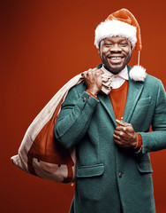 Modern Santa Claus. Smiling emotional man posing in green coat and red sweater, with santa hat and bag. Studio shot, red background