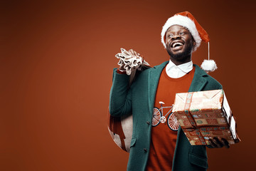 Modern Santa Claus. Smiling emotional man posing in green coat and red sweater, with santa hat and bag and present. Studio shot, brown background - 241211852