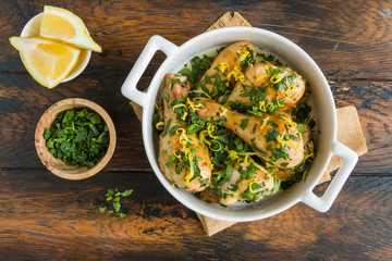 Baked chicken legs with fresh parsley and lemon juice and zest in white casserole on wooden rustic table, top view - 241213631