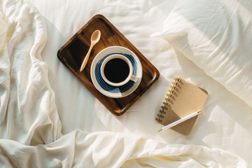Flatlay with cup of coffee or tea on wooden tray with notebook and pen on side on white sheets, selective focus