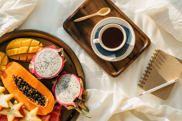 Flatlay with cup of coffee or tea on wooden tray and tropical fruit plate with notebook and pen on side on white sheets, selective focus