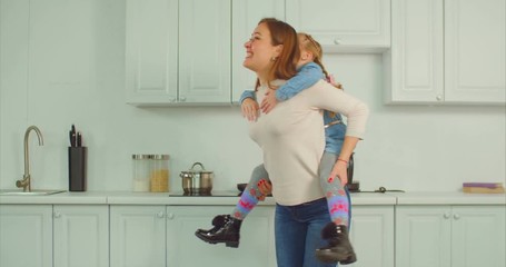 Carefree positive attractive mother giving piggyback ride to excited elementary age daughter with pigtails in domestic kitchen. Joyful cheerful family with little girl playing and having fun indoors.