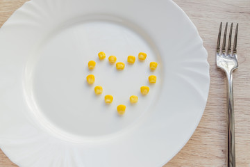 Close up of sweet corn in shape of heart and steel fork on white plate.