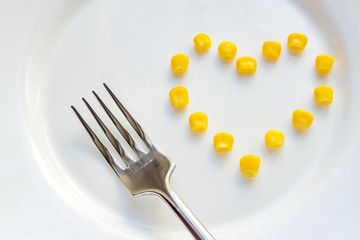 Close up of sweet corn in shape of heart and steel fork on white plate.