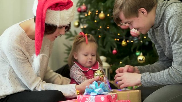 Happy family, parents showing Ney Year decorations to their cute child in front of Christmas tree. Mother in Santa hat, father, and baby girl with Christmas toys and presents near beautiful spruce.