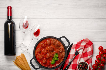 Meatballs in tomato sauce in a frying pan with cherry, tomatoes, bottle of wine and two glasses on a white wooden board