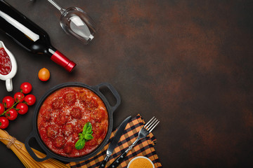 Meatballs in tomato sauce with spices, cherry tomatoes, pasta and basil in a frying pan with bottle of wine and wineglass on rusty brown background