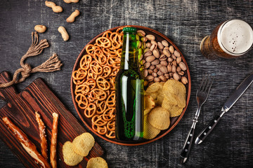 A bottle of beer on a plate with salted ookies pretzels, pistachio nuts and chips on a black scratched chalk board