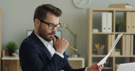 Portrait shot of the Caucasian young handsome businessman in glasses reading and checking some documents or contract in his office room.