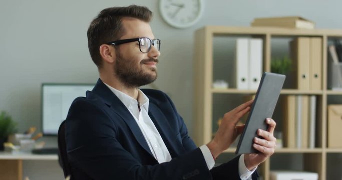 Caucasian young good looking businessman scrolling and tapping on his tablet device, then rising his head and smiling. In his cabinet. Indoors.