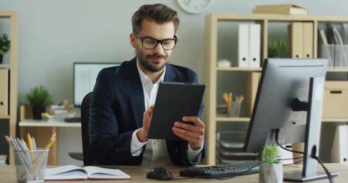 Young handsome Caucasian man scrolling and tapping on the tablet computer in his comfortable office during daytime.