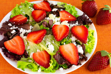 Vegetarian delicious fresh salad with purple and green lettuce leaves, strawberries and goat cheese on orange tablecloth.