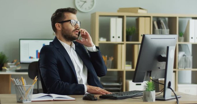 Caucasian young man communicating about his business on the mobile phone while being in his cabinet at the table.