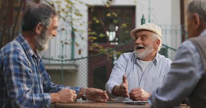 Happy senior Caucasian man in a cap winning a domino game from his neighbor friends in the yard at the table. Outdoor.