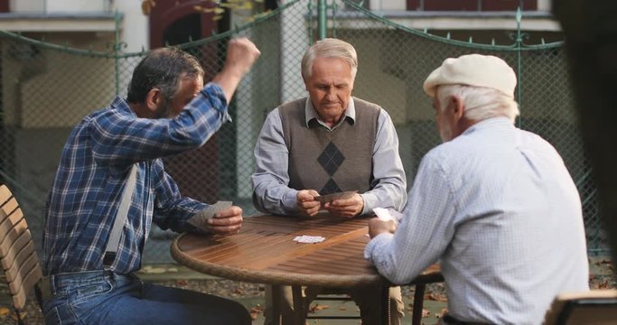 Caucasian old best friends playing cards game at the fresh air and making their turns on the wooden table. Outdoor.