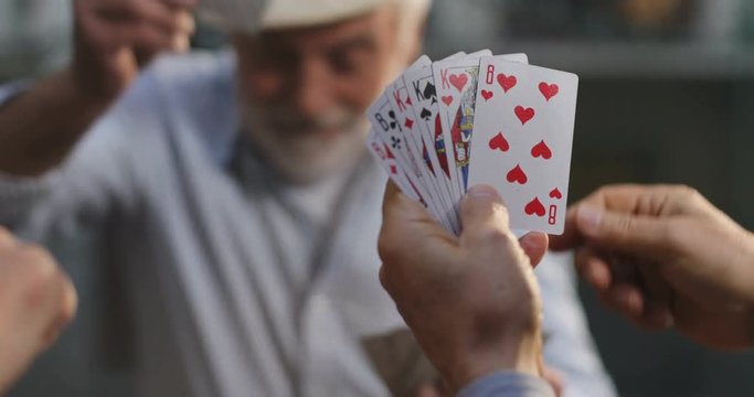 Close up of the play cards in the hands of an old man and he taking one for his turn. Another player on a background. Outside.