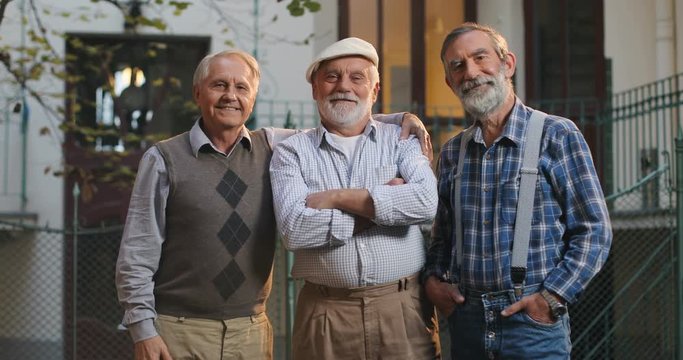 Portrait shot of the three senior Caucasian men friends standing and smiling to the camera in front of the camera in the yard. Outdoors.