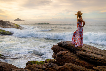 Beautiful slender woman with a hat and stylish dress standing on a beach with the waves crashing into the rocks splashing with the wider seascape at beautiful sunrise