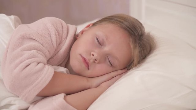 A close-up of long-haired girl in a gown lying sidelong in the bed with her eyes closed and sleeping. She moves her lips slightly. The camera moves from right to left and it freezes on girl's face
