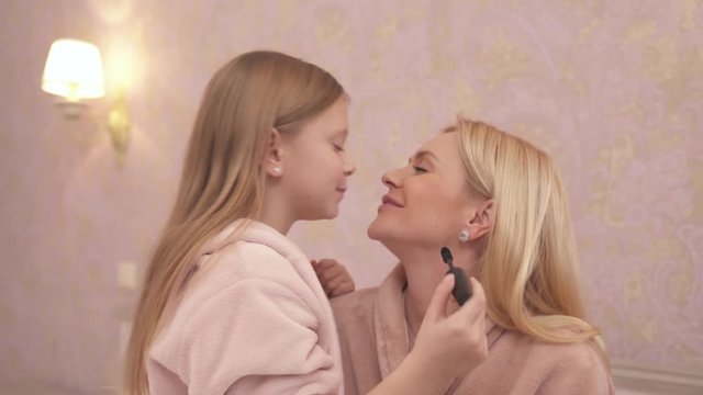 A close-up side view of a woman and a girl in gowns facing each other. The girl is holding mascara. They smile, kiss