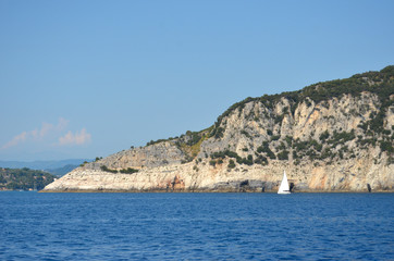 Fototapeta na wymiar A yacht with a white sail is on the blue waters of the Mediterranean Ocean. Behind is a rocky island, dotted with scrub. The mainland with hills is in the distance. The sky is blue.