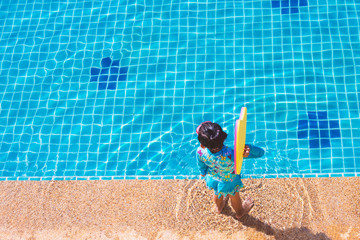 Asian child girl in swimsuit standing and holding floating foam side the pool.