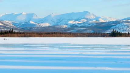 Frozen lake in winter mountains at sunrise. Snowy mountains in the rays of dawn. Winter landscape on the mountains and the frozen lake in Yakutia, Siberia. Shadows in the frozen lake at morning. Pano