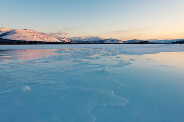 Frozen lake in winter mountains at sunrise. Snowy mountains in the rays of dawn. Winter landscape on the mountains and the frozen lake in Yakutia, Siberia. Shadows in the frozen lake at morning. Pano