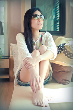 Asian woman wearing sunglasses and looking out the window in vintage style. Portrait of lovely asian young woman dressed in stylish posing while sitting on sofa near terrace.
