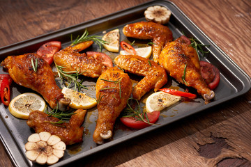 Baked chicken drumstick and wings on baking tray over dark wooden background.