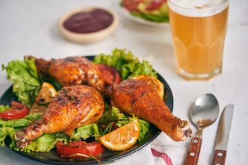 Grilled chicken legs roasted on the grill on dark plate with tomato sauce in a bowl and lettuce leaves, glass mug of beer