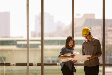Two Asian engineer, male with yellow safety helmet and woman with white one standing and talking near high curtain wall glass frame in construction site