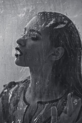 Black and white shot of model with wet hair dressed in black swimming suit and transparent rain coat is posing behind the wet transparent membrane in the studio with lighting simulating evening street