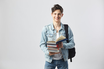 Smiling young dark-haired guy with black backpack  on his shoulder dressed in a white t-shirt, jeans and a denim jacket holds books in his hands on the white background  in the studio