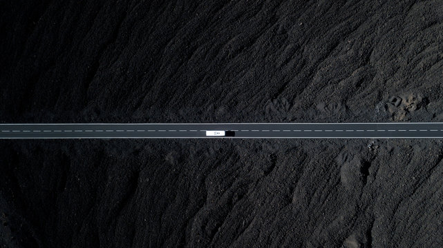 The road with white lines is surrounded by black volcanic lava. Sticking rocks. Dangerous adventure of incredible beauty. Top view, drone footage. Minimalistic landscape. White car in motion.