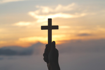 Woman hand holding holy lift of christian cross with light sunset background.