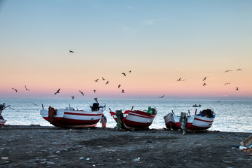 Seagulls flying over boats stranded on the beach with a dramatic background sunset, in Oued Lao, a small fishing village on the coast of the province of Chefchaouen, Morocco