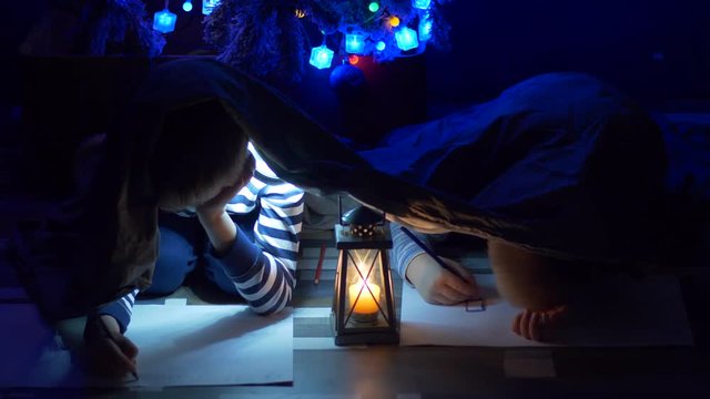 Funny children write letter with pictures of Santa Claus under fir, covered with blanket and flashlight. Children are happy to spend winter holidays together. Concept of traditional family holidays.
