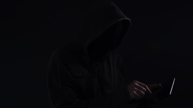 Hooded computer hacker with obscured face using digital tablet in cybercrime and cybersecurity concept, low key slow motion