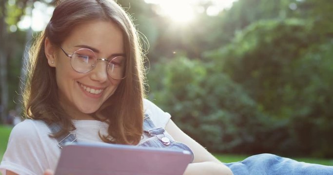Close up of the beautiful young Caucasian woman watching something on the tablet computer while lying and resting in the park.