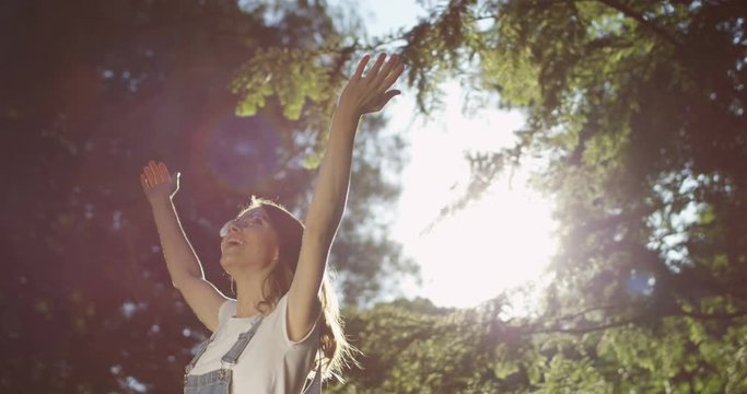 Portrait shot of the joyful Caucasianh girl in glasses having fun while whirling with hands up in the bautiful park on a sunny summer day.