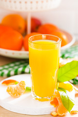 Obraz na płótnie Canvas Tangerine orange juice in glass and fresh fruits with leaves on white wooden kitchen background closeup. Healthy and tasty refreshing summer beverage